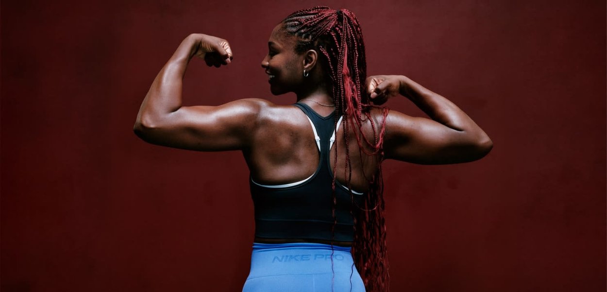 Donna atletica con lunghe treccine rosse che mostra i muscoli delle braccia, indossando un top sportivo nero e leggings Nike Pro blu, su sfondo marrone scuro.