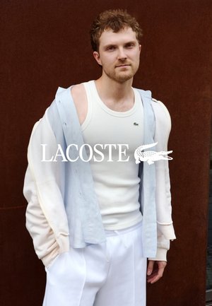 Junger Mann trägt weißes Tanktop, hellblaues Hemd und cremefarbene Jacke, steht vor dunkelbraunem Hintergrund, Lacoste-Logo auf der Brust sichtbar.