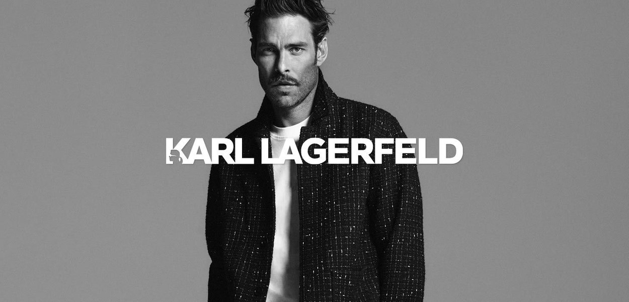 Blazer noir structuré sur une chemise blanche, mannequin avec des cheveux ébouriffés et une barbe, sur fond gris. Nom de la marque affiché de manière proéminente.