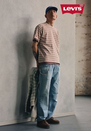 Junger Mann mit gestreiftem Hemd, hellblauen zerrissenen Jeans mit Kette, der eine Jacke hält und vor einer Wand mit dem Levi's-Logo darüber steht.