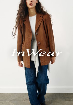 Blazer en daim marron avec un col cranté, deux boutons et des poches avant, associé à un haut en couches gris clair et un jeans bleu à jambes larges.