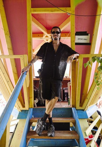 Schwarzes, mesh-artiges Hemd mit Knöpfen, ausgefranste schwarze Shorts und graue Sneakers. Holztreppen mit blauen und gelben Akzenten, pinke Wände, sichtbares "NUR FÜR KÜNSTLER"-Schild.