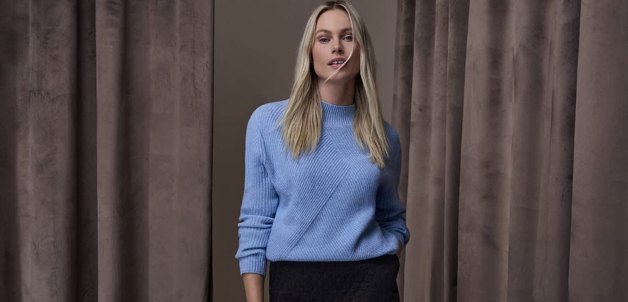 Pull bleu clair en maille avec texture côtelée et détails de coutures diagonales, assorti à une jupe foncée. En arrière-plan, des rideaux marron.