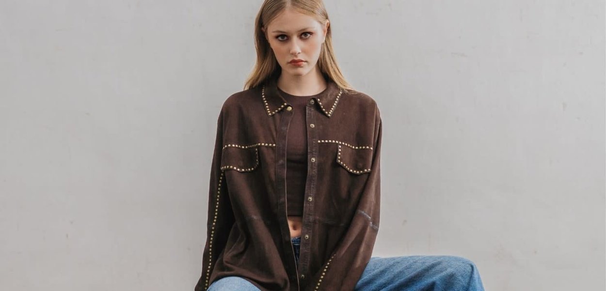 Giovane donna con lunghi capelli biondi che indossa una maglia marrone scura con borchie e jeans blu, seduta contro un muro semplice di colore grigio chiaro.