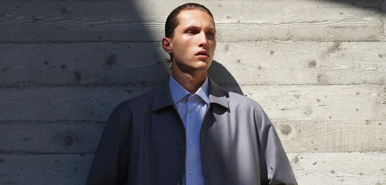 Giacca leggera grigia con una morbida texture, indossata sopra una camicia bianca con colletto, su uno sfondo di muro di cemento testurizzato.