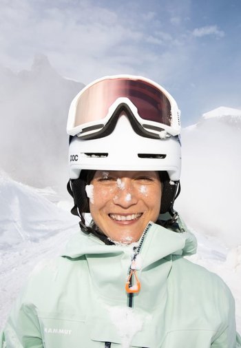 Weißer Skihelm mit Visier, mintgrüne Jacke, Schnee im Gesicht und Hintergrund mit schneebedeckter Landschaft und Wolken. Helle und strukturierte Details.