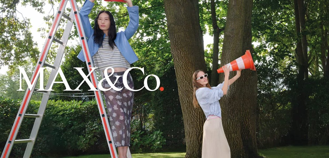 Dwie kobiety na zewnątrz; jedna stoi na drabinie, trzymając górę, druga nosi okulary przeciwsłoneczne i trzyma pomarańczowy stożek drogowy przy ustach.