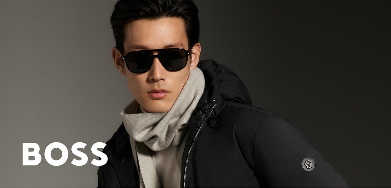 Zwarte gewatteerde jas met capuchon, grijze sjaal en donkere zonnebril. Heeft een cirkelvormig logo op de mouw. Effenkleurig, modern ontwerp.