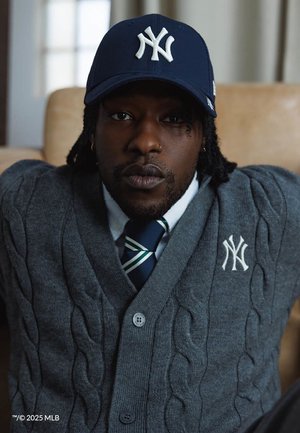 Mand iført mørkeblå New York Yankees kasket, grå kabelstrikket cardigan med Yankees-logo, hvid skjorte og stribet slips, siddende indendørs.