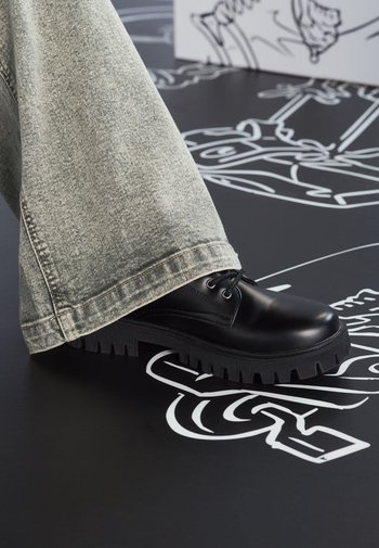Schwarze Schnürschuhe mit einer klobigen, strukturierten Sohle, getragen mit hellgrauen, weiten Jeans mit fransigem Saum.