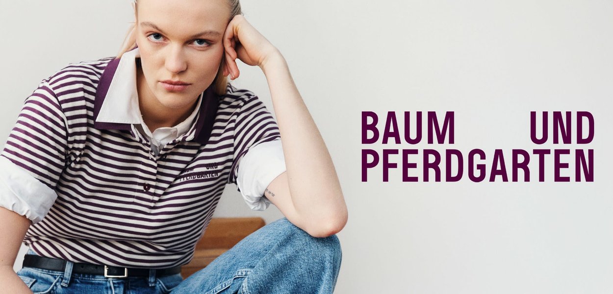 Polo a righe con colletto viola, maniche corte e polsini bianchi, abbinato a jeans a vita alta. Testo "BAUM UND PFERDGARTEN" visibile.