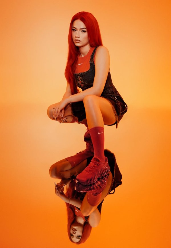 Rote High-Top-Sneaker, rote Kniestrümpfe, schwarze Jeans-Überalls und ein oranges Oberteil, vor einem orangefarbenen Hintergrund mit einer reflektierenden Oberfläche.