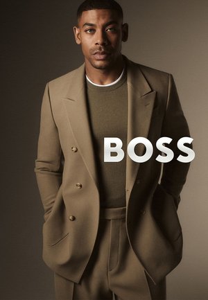 Abito sartoriale doppiopetto beige con maglione verde oliva con trama sotto. Il modello posa con le mani nelle tasche su uno sfondo neutro.