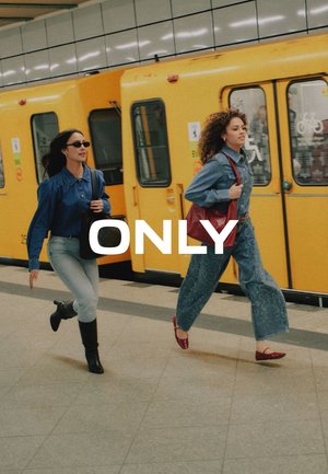 Due donne che camminano rapidamente su una banchina della metropolitana accanto a un treno giallo; una indossa occhiali da sole, l'altra porta una borsa rossa.