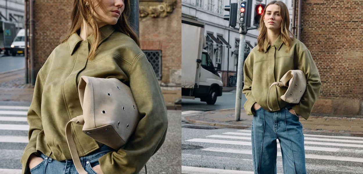 Junge Frau steht an einem städtischen Zebrastreifen, trägt eine olivgrüne Jacke und blaue Jeans und hält eine beigetönte Tasche mit Metallösen unter dem Arm.