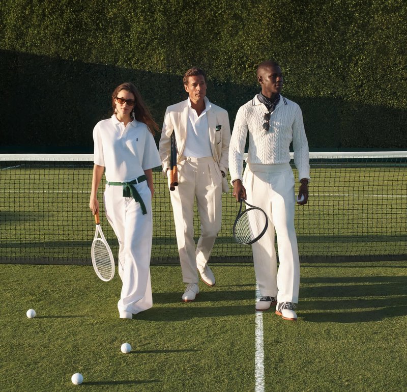 Tres personas vestidas de blanco caminan por una pista de tenis de césped sosteniendo raquetas, con pelotas de tenis esparcidas cerca de la red detrás de ellos.