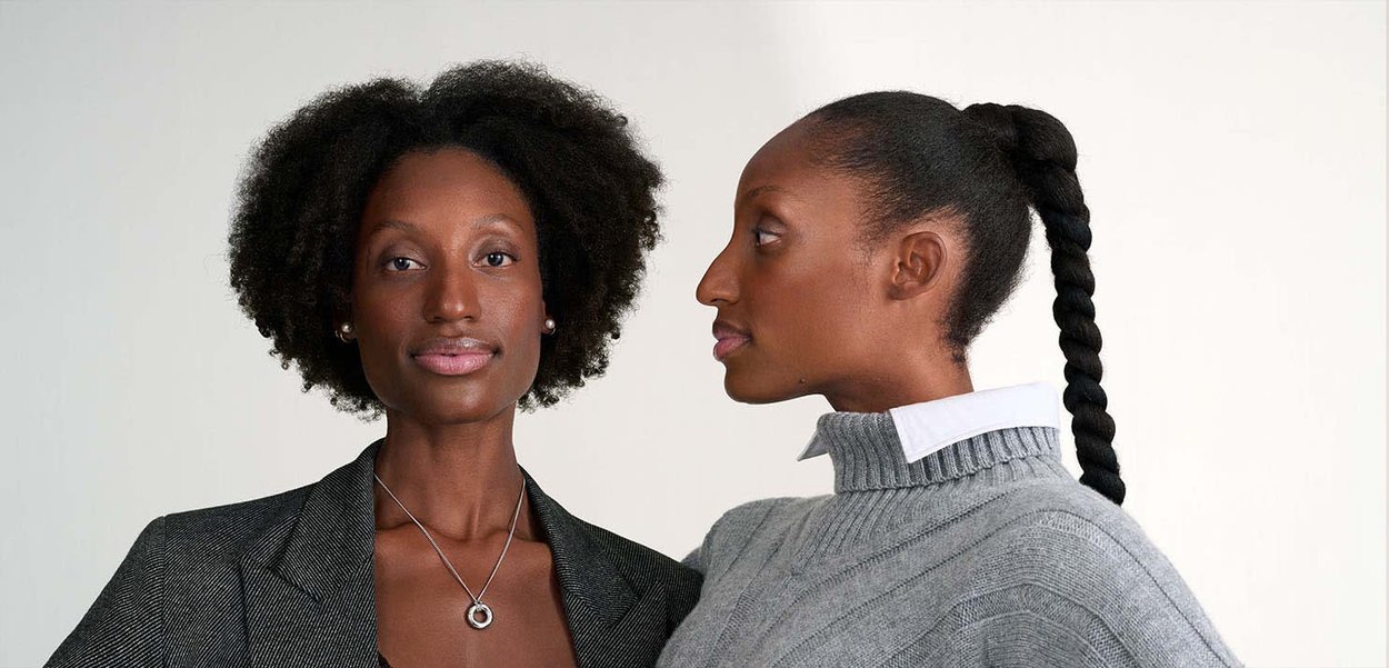 Due donne sono mostrate. La sinistra indossa un blazer scuro e testurizzato e una collana. La destra ha un maglione grigio chiaro con un colletto a coste e una treccia.