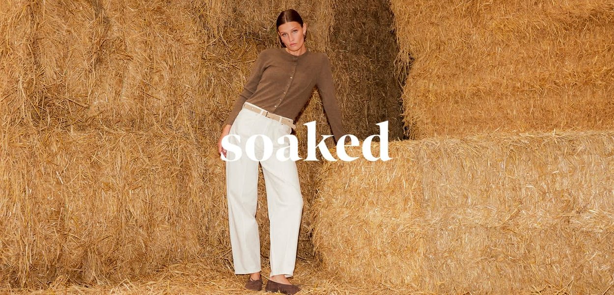Frau lehnt an gestapelten Heuballen, trägt braunen Pullover, weiße Hose und braune Schuhe, mit dem Wort „soaked“ in weißer Schrift darübergelegt.