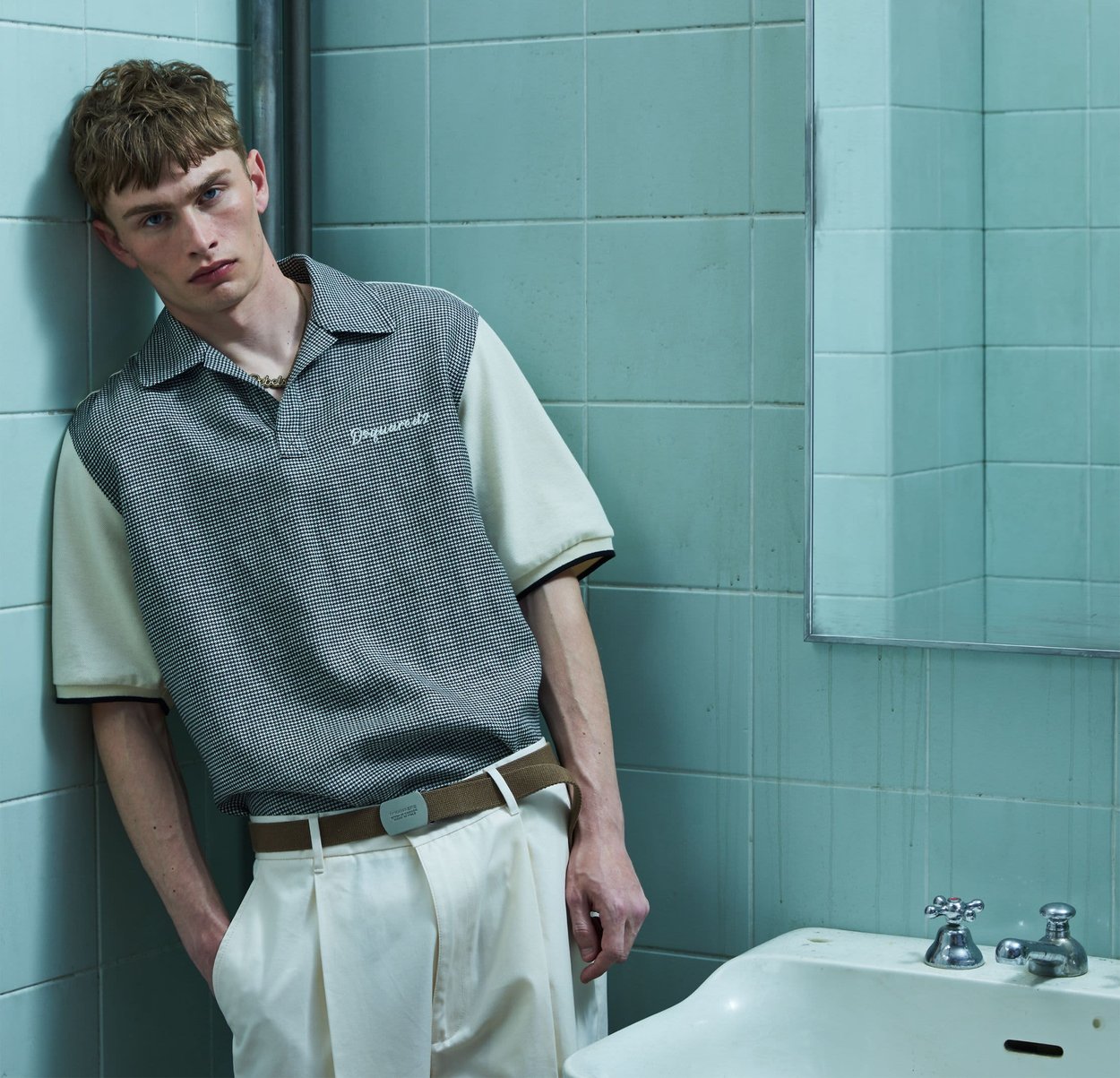 Joven con pantalones beige y camisa polo gris con patrón, apoyado contra una pared de azulejos verdes claros junto a un lavabo blanco con grifos plateados.