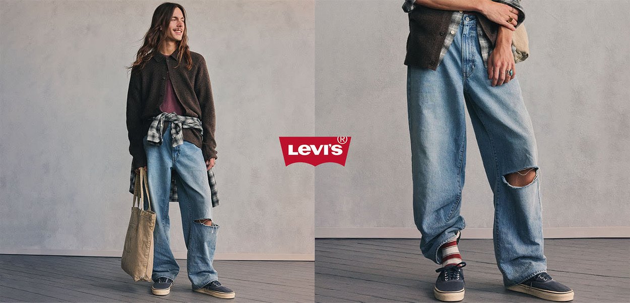 Junge Person trägt übergroße zerrissene Jeans, braune Jacke, gestreifte Socken und Turnschuhe, hält eine beige Tragetasche mit zentriertem Levi's-Logo.