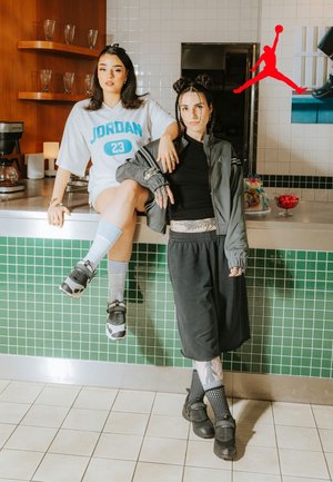 Two individuals, one in a white oversized jersey and the other in a black top, wearing black chunky shoes, stand on a tiled surface.