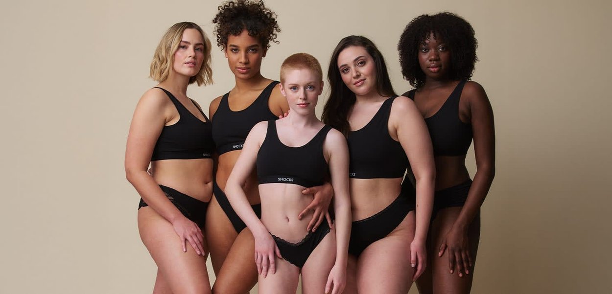 Fem forskellige kvinder står tæt sammen iført matchende sort undertøjssæt mod en neutral beige baggrund.