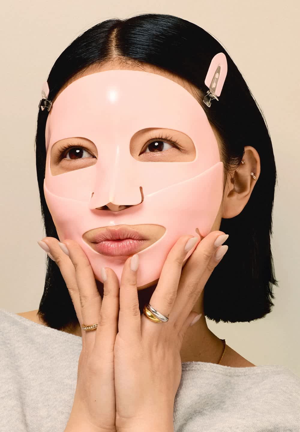 Vrouw met zwart haar die een roze gezichtsmasker draagt dat met clips is bevestigd, haar wangen aanraakend met handen waarop gouden en zilveren ringen zitten.
