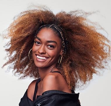 Portrait of a person with voluminous, textured hair in shades of brown and blonde, featuring a braided section, wearing hoop earrings and a black top.