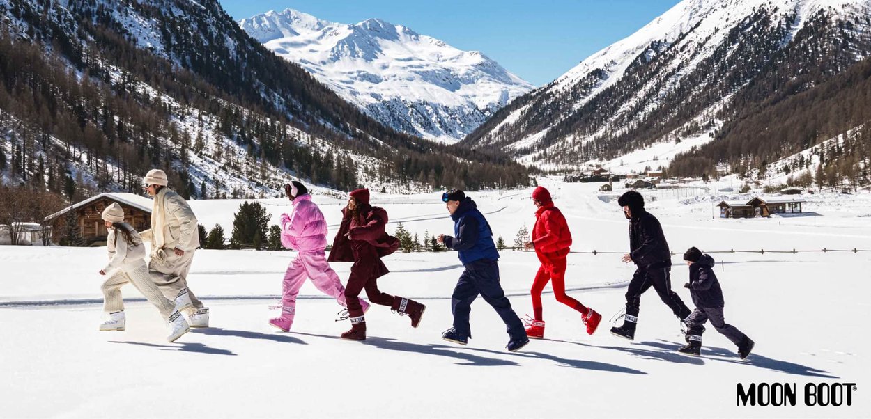 Sette persone indossano abbigliamento invernale colorato e Moon Boots, correndo attraverso un paesaggio innevato con montagne sullo sfondo.