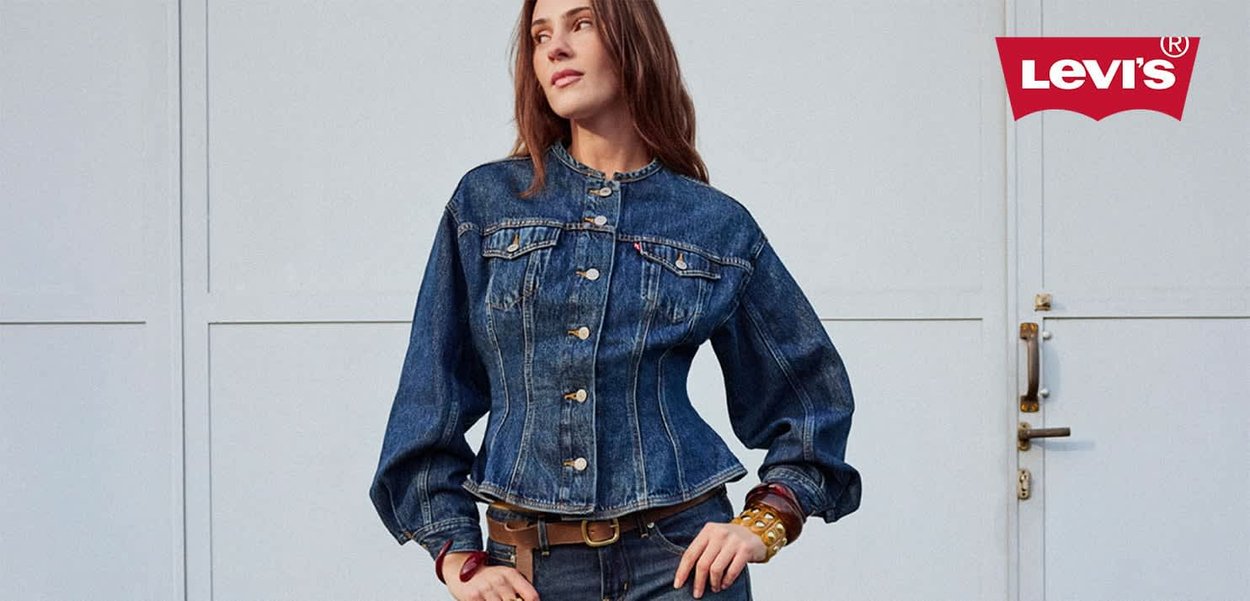 Frau in einer taillierten blauen Jeansjacke und Jeans steht mit den Händen in die Hüften gestützt vor einer hellen Wand, Levi's-Logo rechts oben.
