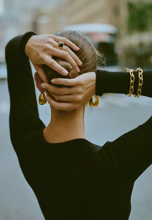 Boucles d'oreilles dorées, bracelet en chaîne épaisse, pull noir à côtes. Mains dans les cheveux, texture et détails des bijoux mis en valeur.