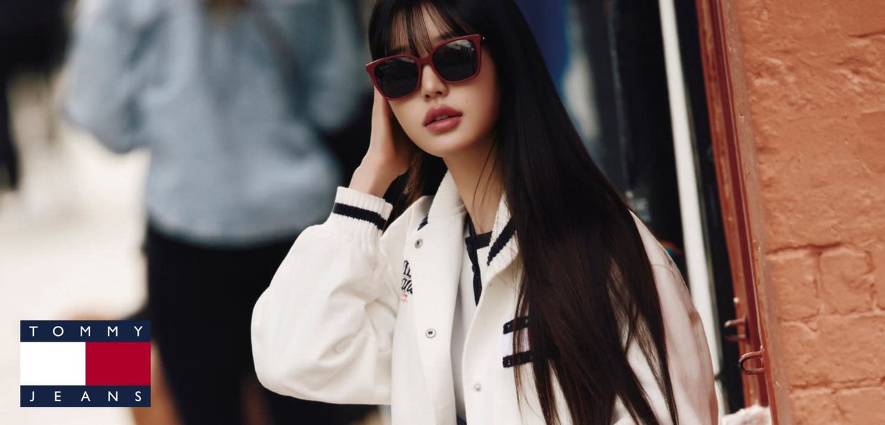 Młoda kobieta z długimi ciemnymi włosami, w czerwonych okularach przeciwsłonecznych i białej kurtce varsity, stoi na zewnątrz obok muru z cegieł.