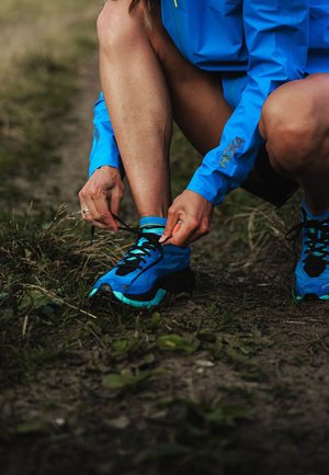 Blue running shoes with black accents, textured material, and thick soles. Laces are being tied, with grass and dirt visible in the surroundings.