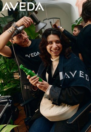 Aveda-handdoek met merklogo, groene haarproducten in de hand, stoel op de voorgrond, professionele haardroger erboven gehouden, omringd door groen en mensen.