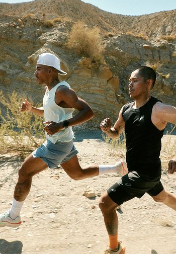 Two male runners in an outdoor environment. One wears a light blue sleeveless shirt and shorts; the other has a black sleeveless shirt and shorts.