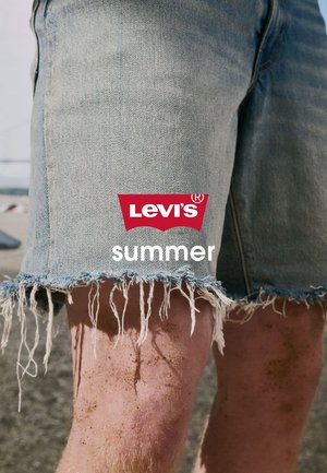 Shorts di denim azzurro chiaro sfilacciati indossati su gambe sabbiose in spiaggia con il logo rosso Levi's e la parola "summer" sovrapposta.