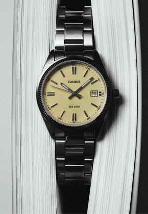 Casio-Armbanduhr mit beigem Zifferblatt, schwarzem Metallarmband, Datumsanzeige zeigt 18, liegend zwischen den Seiten eines aufgeschlagenen Buches.
