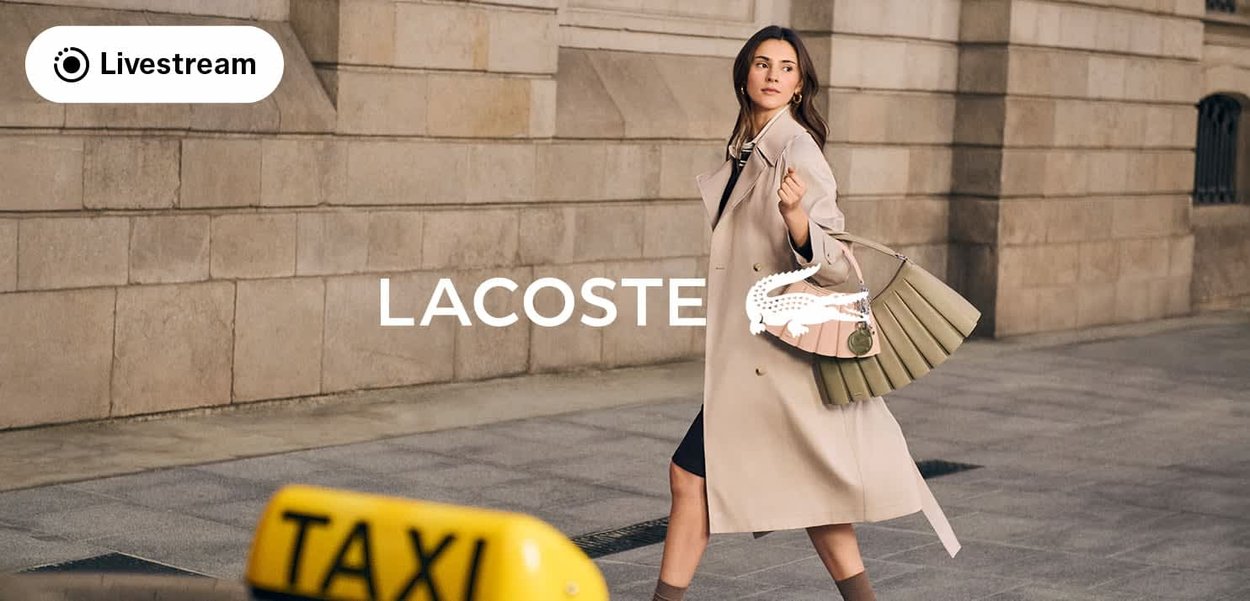 Mujer con abrigo beige y bolso plisado caminando por la calle de la ciudad con un cartel de taxi, el logo de Lacoste y un icono de transmisión en vivo visibles.