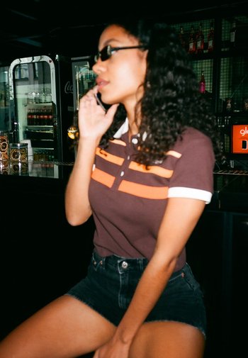 Bruine polo met oranje strepen, korte mouwen en een kraag, gecombineerd met hoge zwarte denimshorts. Donker, golvend haar.