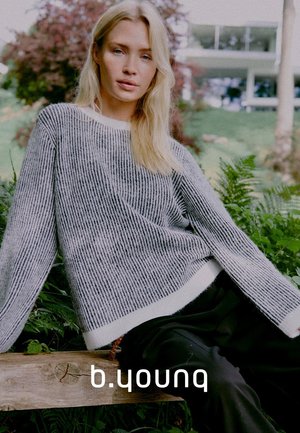 Grau-weiß gestreifter Pullover mit lockerer Passform und strukturiertem Strick, kombiniert mit schwarzen Hosen. Grüne Pflanzen im Hintergrund.