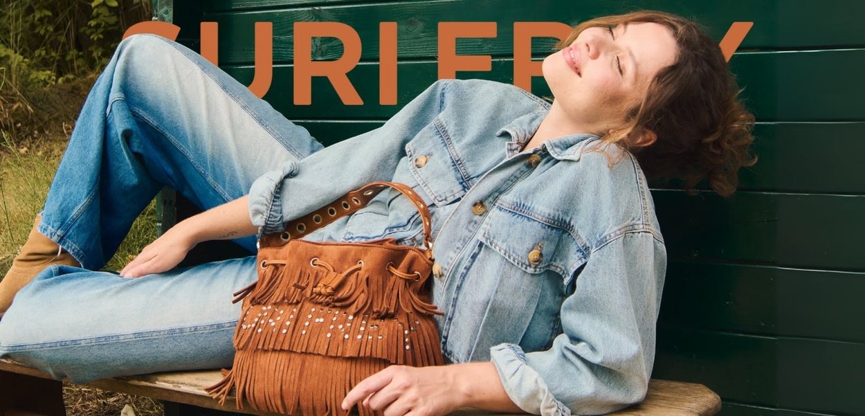Brown suede bucket bag with fringes and silver studs, worn over a denim jacket. Model is resting against a green wooden background.