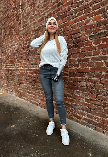 Vit kabelstickad tröja, grå tajta jeans och vita sneakers. Har en silverfärgad protesarm med svart hårdvara, mot en tegelvägg.
