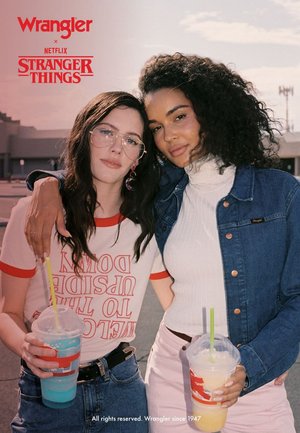 To personer står tett sammen, iført en hvit ribbestrikket turtleneck, en denimjakke og en grafisk t-skjorte med røde detaljer. Begge holder fargerike drikker.