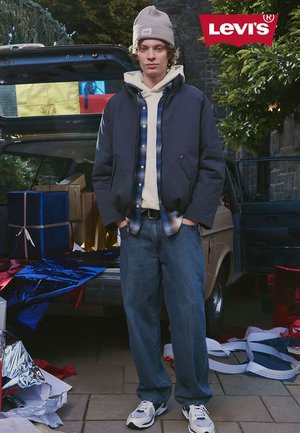 Levi's jacket in navy with gray accents, layered over a beige hoodie and plaid shirt, paired with blue jeans and white sneakers.