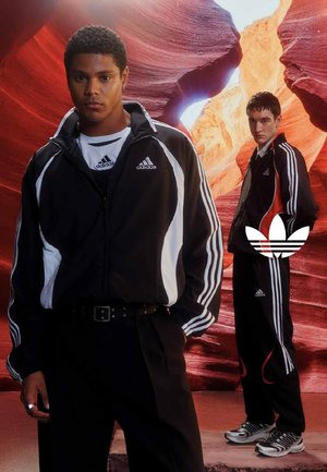 Zwei Männer in schwarzen Adidas-Trainingsanzügen stehen in einem roten Canyon, durch den Sonnenlicht durch Felsformationen fällt.