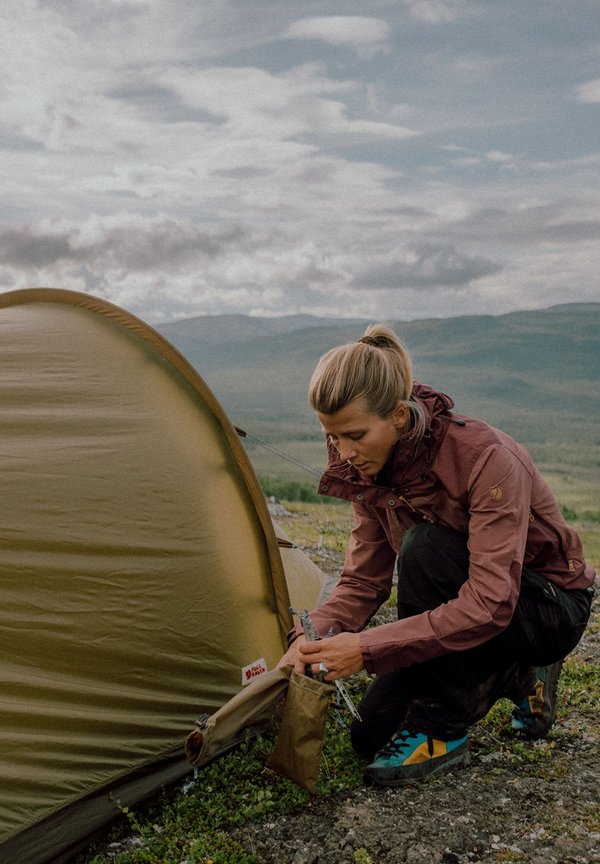 Tält med en rundad, olivgrön design; person i en vinröd jacka och svarta byxor fäster det med metallspikar på stenig mark.