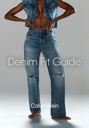 Person wearing ripped blue denim jeans and a cropped denim vest, standing barefoot against a plain gray background.