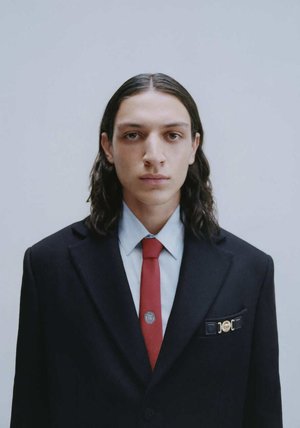 Abito in tessuto nero, camicia blu e cravatta rossa. Hardware dorato notevole sulla tasca sinistra. I capelli sono lunghi e scuri. Sfondo neutro.