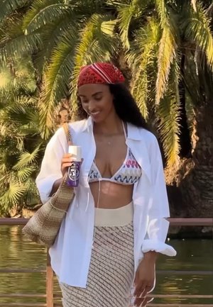 Frau in gemustertem Bikini-Oberteil und cremefarbener Rock hält eine lila Flasche nahe Wasser mit Palmen im Hintergrund, lächelt und schaut nach unten.