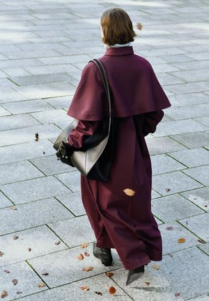 Abrigo largo color granate con diseño de capa superpuesta, hecho de tela texturizada. Bolso negro para el hombro con forma redondeada, llevado en el brazo izquierdo.